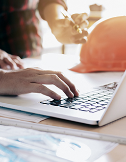 Mann schreibt auf Laptop, im Hintergrund liegt ein Arbeitshelm auf dem Tisch